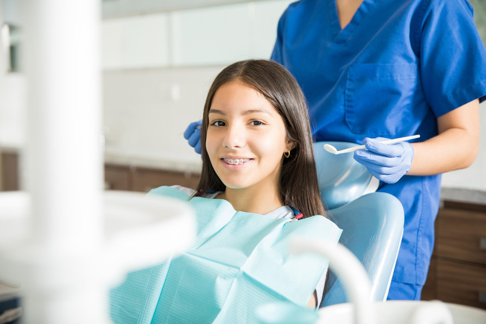 A young teenage girl with braces smiling and sitting on a dentist chair while the dentist is behind in the background