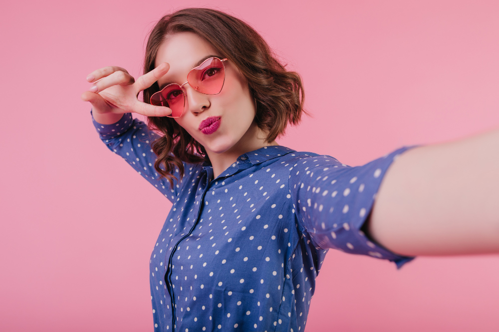 A woman pouting her lips in a blue dotted shirt taking a selfie with a pink background